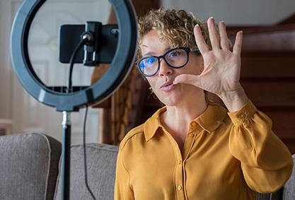 Image shows a woman in a yellow shirt with her hand up as she records a video.
